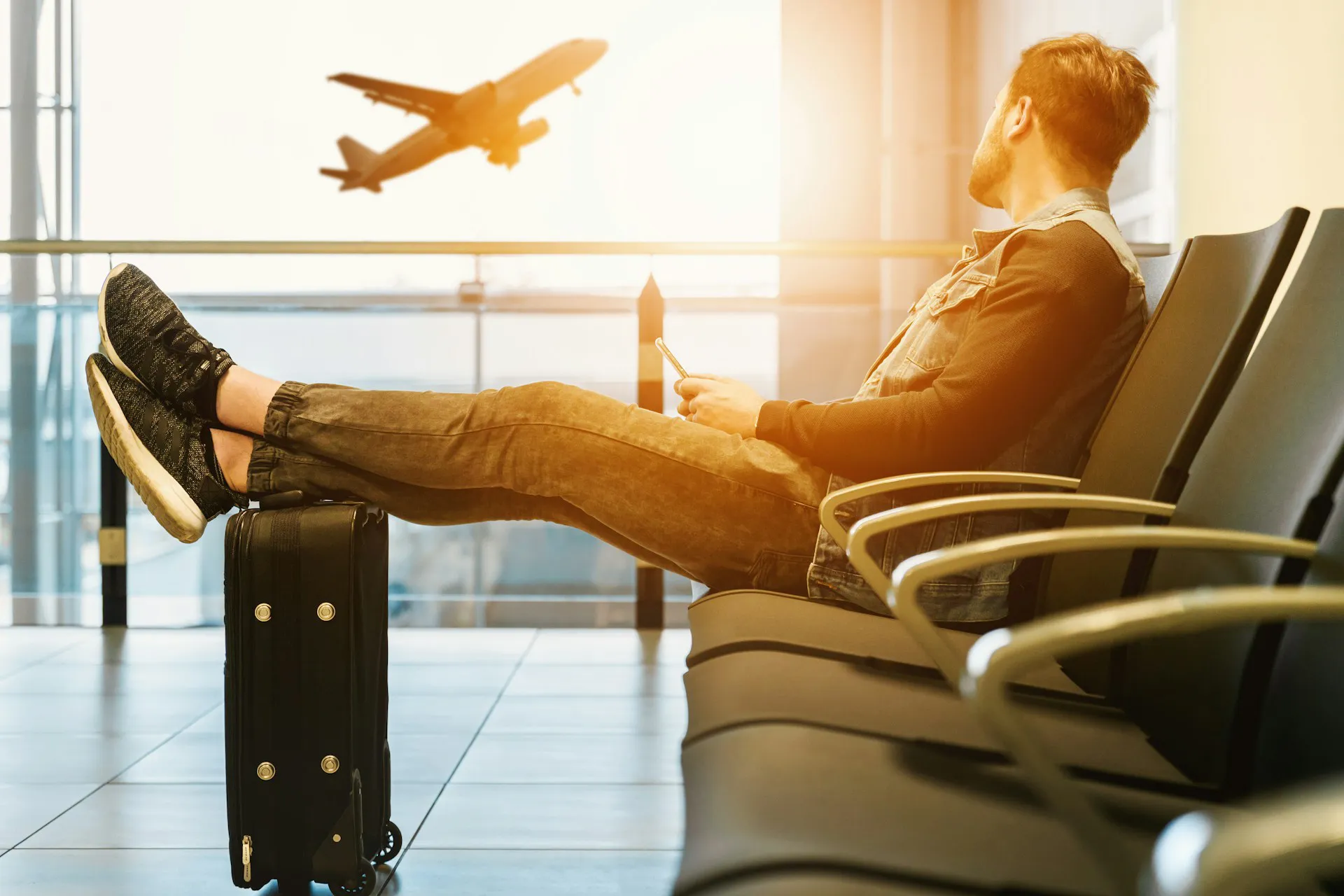 Un homme assis à l'aéroport avec ses pieds posés sur sa valise, regardant un avion qui décolle